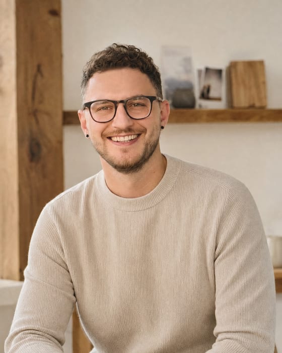 Ein Mann mit Brille, kurzen dunklen Locken und hellem Strickpullover sitzt lächelnd in einem hellen Raum. Hinter ihm ein Holzbalken und ein Wandregal mit Fotos.